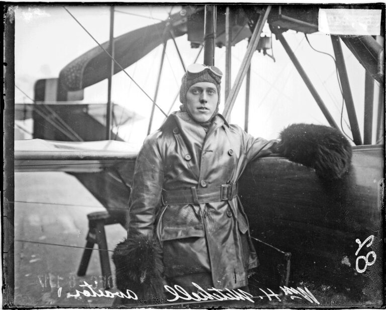 Black & white photograph of a man standing next to an airplane that is resting on sawhorses. He is wearing an aviator cap and goggles atop his head, a coat, and big fur mitts on his hands.