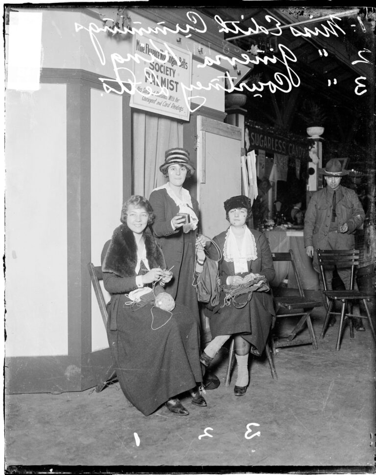 Black & white photograph of three young white women--two are sitting and holding yarn and knitting needles, the one in the middle is standing.