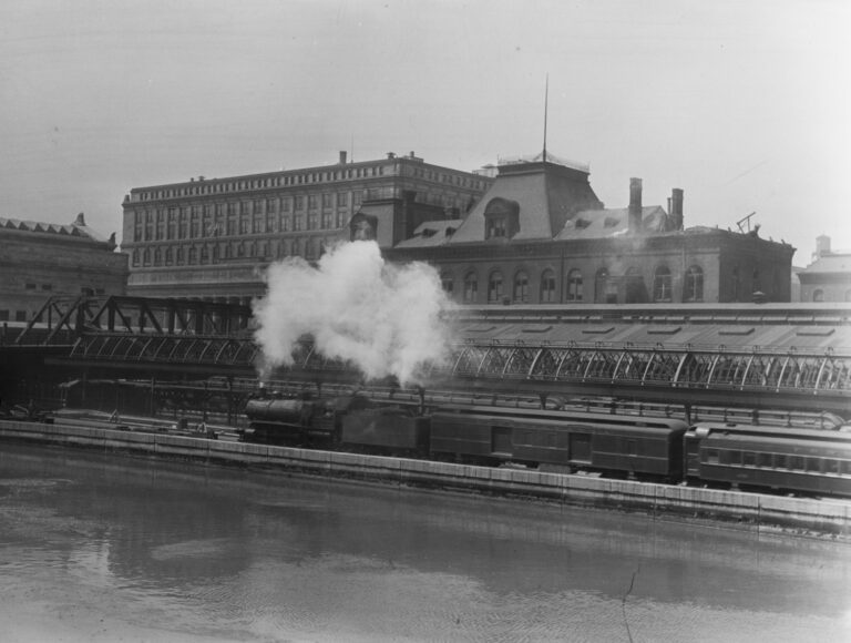 Black & white photograph of old and new train station buildings. A steam engine with passenger cars is passing in the foreground, next to a body of water. 